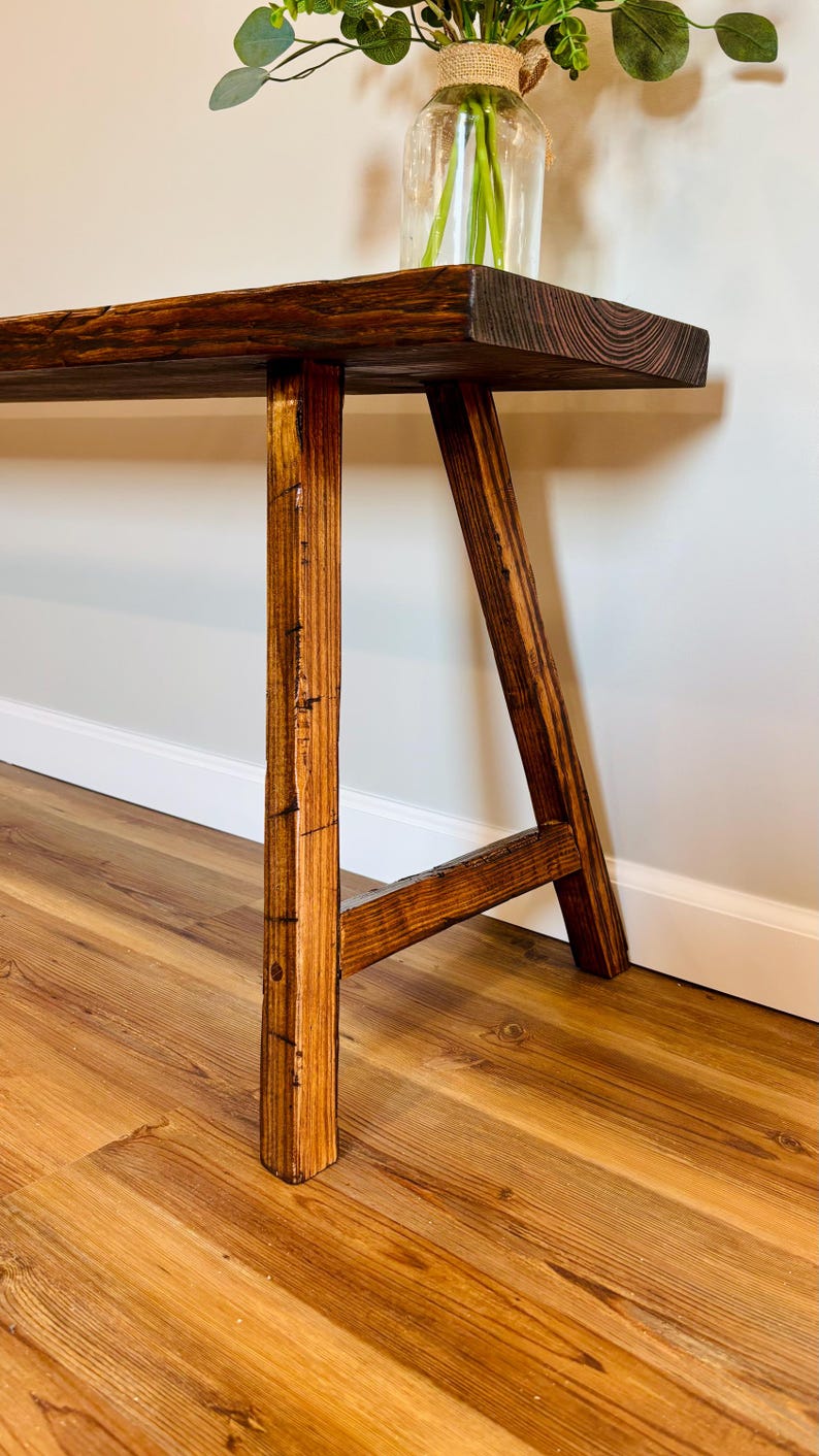 May include: A rustic wooden bench with a dark stained top and light brown legs. The bench has a natural wood grain and a simple, sturdy design. A glass vase with greenery sits on top.
