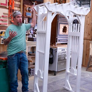 May include: A white wooden arbor with an arched top and vertical slats. The arbor is constructed with a series of posts and beams, creating a decorative garden structure. The arbor is designed to support climbing plants or serve as a decorative entrance.