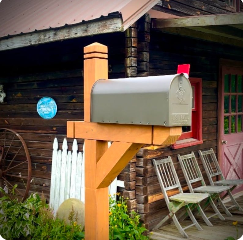 May include: A gray mailbox with a wooden post mounted on a wooden porch of a rustic log cabin. The mailbox has a flag and the text "US Mail" on it.