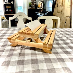 May include: A wooden paper towel holder shaped like a miniature picnic table. The holder is made of natural wood and has a paper towel roll inserted into the table's center.