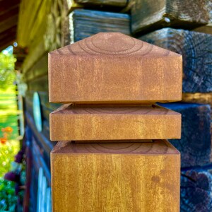 May include: A close-up of a wooden fence post with a layered, pyramid-shaped top. The post is stained a rich brown color.