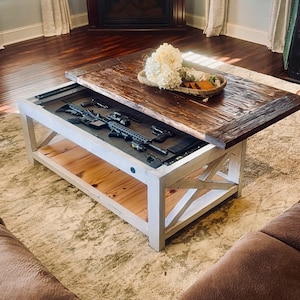 May include: A white farmhouse style coffee table with a hidden compartment that opens to reveal a gun safe. The table top is made of dark wood and has a distressed finish. The table is sitting on a beige rug.