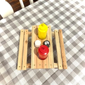 May include: A wooden picnic table with three condiment holders. The holders are filled with a yellow mustard bottle, a red ketchup bottle, and a white salt shaker. The table is on a gray and white checkered tablecloth.