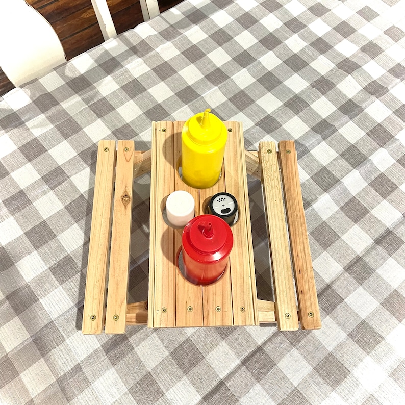 May include: A wooden picnic table with three condiment holders. The holders are filled with a yellow mustard bottle, a red ketchup bottle, and a white salt shaker. The table is on a gray and white checkered tablecloth.