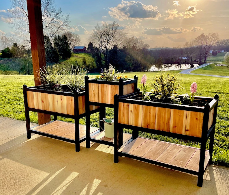 May include: Three wooden planter boxes with black metal frames and legs. The planters are filled with various plants and flowers. The planters are on a patio with a view of a green lawn and a pond in the background.