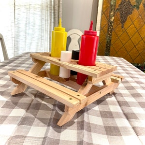 May include: A miniature wooden picnic table condiment holder with a yellow mustard bottle, a red ketchup bottle, and two white condiment bottles.