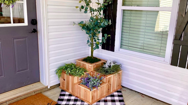 May include: A wooden planter with four compartments, each containing a different type of plant. The planter is in a corner of a porch, with a white door and a window to the right.