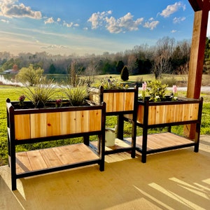 May include: Three wooden planter boxes with black metal stands. The planters are filled with plants and flowers. The planters are on a patio with a view of a lake and trees in the background.