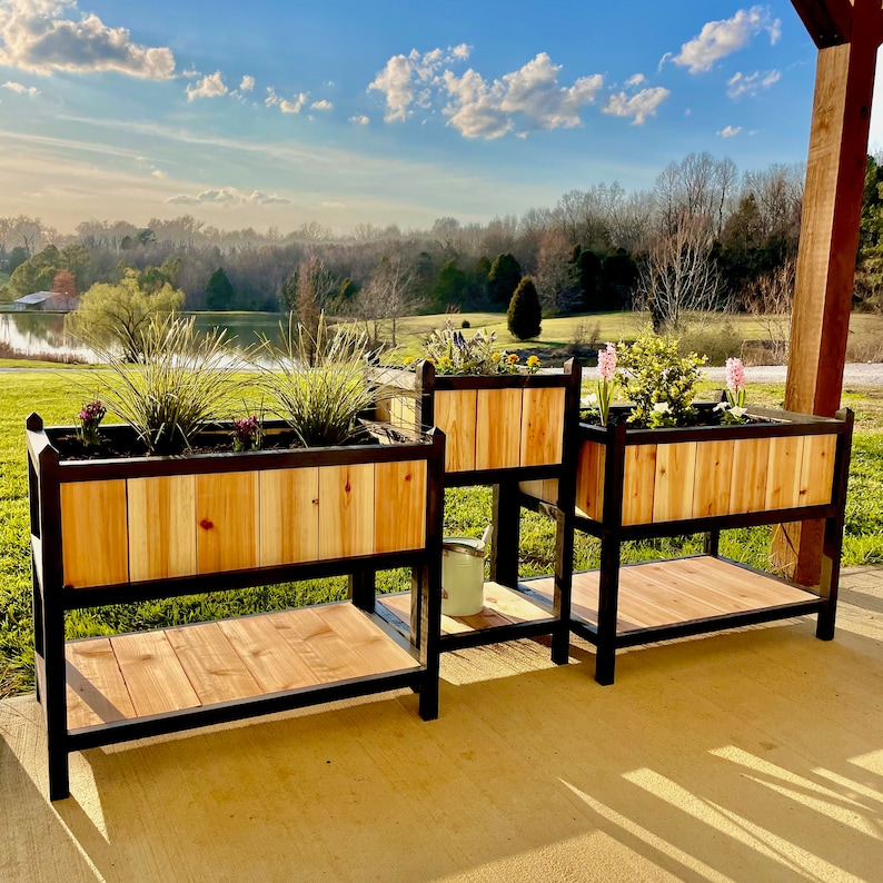 May include: Three wooden planter boxes with black metal stands. The planters are filled with plants and flowers. The planters are on a patio with a view of a lake and trees in the background.