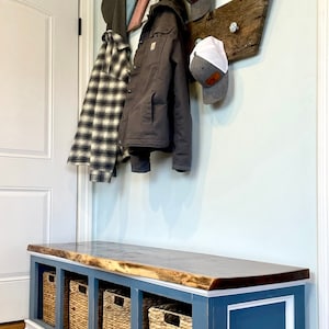 May include: A blue painted bench with a live edge wood top and four woven baskets. The bench has white trim and is against a light blue wall with a wooden arrow and hats hanging on it. The sign above the door says "Laundry".
