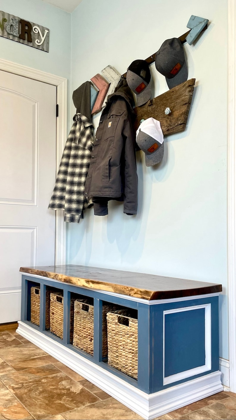 May include: A blue painted bench with a live edge wood top and four woven baskets. The bench has white trim and is against a light blue wall with a wooden arrow and hats hanging on it. The sign above the door says "Laundry".