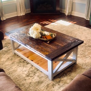 May include: A rustic wooden coffee table with a white painted base and a lower shelf. The table has a distressed wood finish and features a white X-shaped design on the base. A bouquet of white flowers is sitting on the table.