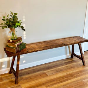 May include: A rustic wooden bench with a natural finish. The bench has a long, rectangular seat supported by angled legs. A small bouquet of greenery and several books are placed on the bench, adding a touch of decor. The bench is set against a neutral wall.