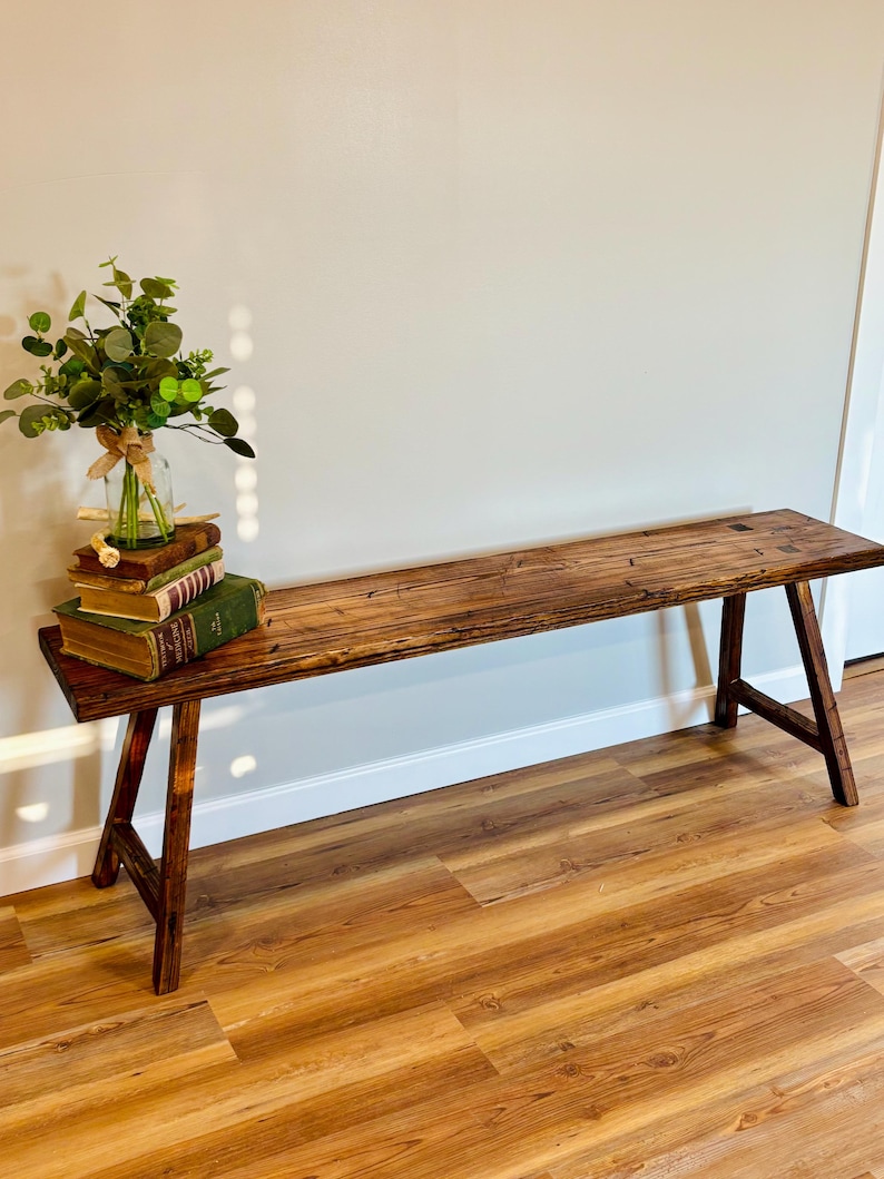 May include: A rustic wooden bench with a natural finish. The bench has a long, rectangular seat supported by angled legs. A small bouquet of greenery and several books are placed on the bench, adding a touch of decor. The bench is set against a neutral wall.