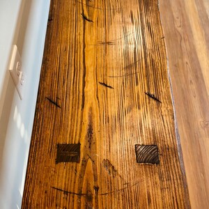May include: A close-up view of a handcrafted wooden table. The table has a warm, natural wood tone with dark grain patterns and visible knots. Two square wooden inlays are visible. The table's surface is textured and appears to be well-finished.