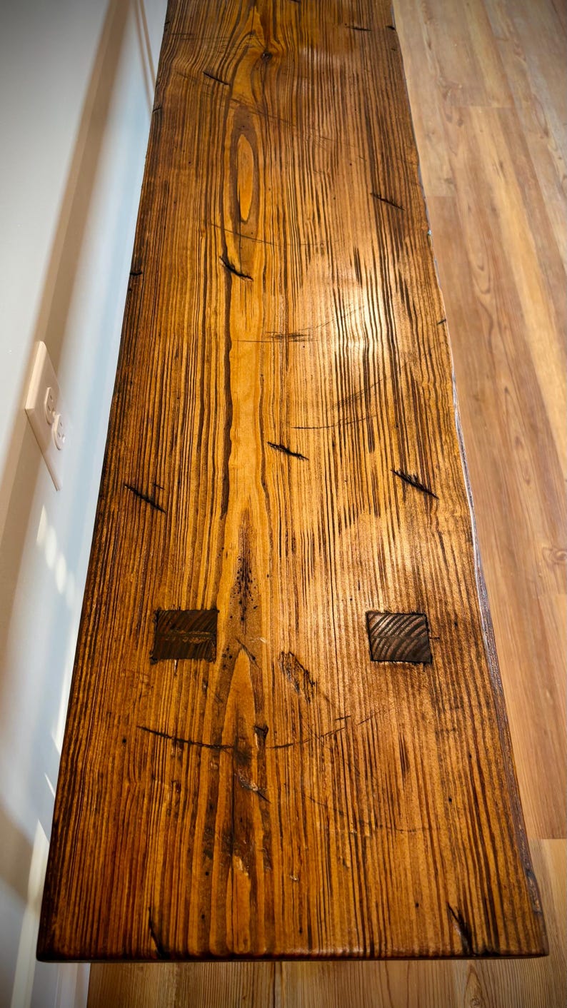 May include: A close-up view of a handcrafted wooden table. The table has a warm, natural wood tone with dark grain patterns and visible knots. Two square wooden inlays are visible. The table's surface is textured and appears to be well-finished.