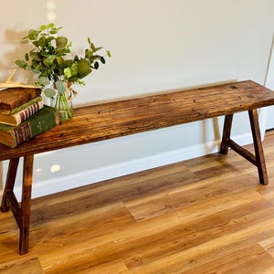 May include: A rustic wooden bench with a dark stain finish. The bench has a rectangular top and angled legs. A small arrangement of books, greenery, and a decorative antler sits on the bench. The bench is approximately 60 inches long.