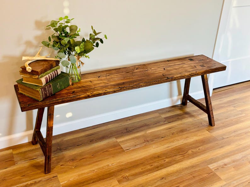 May include: A rustic wooden bench with a dark stain finish. The bench has a rectangular top and angled legs. A small arrangement of books, greenery, and a decorative antler sits on the bench. The bench is approximately 60 inches long.