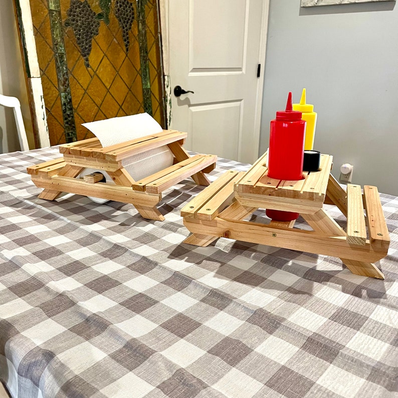 May include: Two miniature wooden picnic tables, one with a paper towel roll and the other with a red and yellow condiment bottle set. The tables are on a checkered tablecloth.