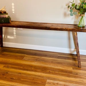May include: A rustic, wooden bench with a dark brown finish. The bench has a rectangular top and angled legs. A stack of books and a vase of greenery sit on the bench. The bench is approximately 60 inches long.