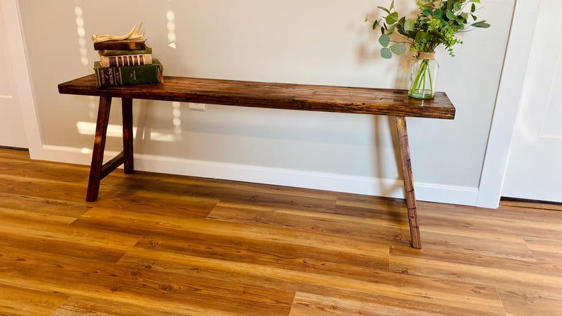 May include: A rustic, wooden bench with a dark brown finish. The bench has a rectangular top and angled legs. A stack of books and a vase of greenery sit on the bench. The bench is approximately 60 inches long.