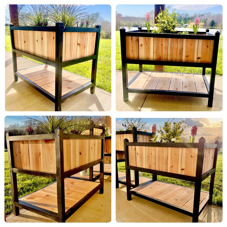 May include: Four black metal plant stands with two shelves. Each stand has a wooden planter box with a black metal frame. The planter boxes are filled with green plants and flowers.
