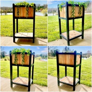 May include: A black metal plant stand with two shelves and a wooden planter box. The stand is in a garden setting with green grass and trees in the background.