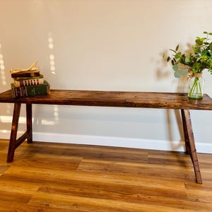 May include: A rustic, dark brown wooden bench with angled legs. The bench is adorned with a stack of vintage books and a glass vase filled with greenery. The bench is placed against a neutral wall and sits on a wood-look floor.
