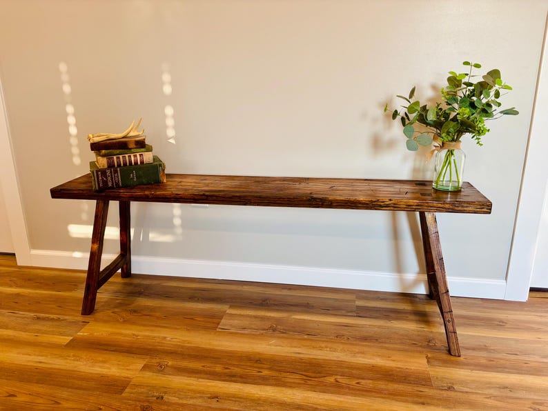 May include: A rustic, dark brown wooden bench with angled legs. The bench is adorned with a stack of vintage books and a glass vase filled with greenery. The bench is placed against a neutral wall and sits on a wood-look floor.