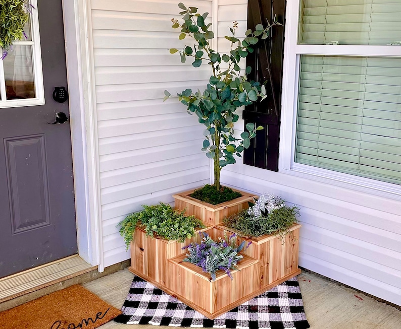May include: A wooden tiered planter with four sections, each containing different plants. The planter is made of natural wood and has a rustic look. The plants include a small tree, green leafy plants, and purple flowers.