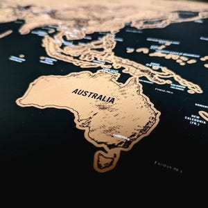 May include: A scratch-off world map featuring a matte black background with metallic gold foil continents and islands. The map displays the continent of Australia and surrounding Pacific islands with clear black text labels for geographic locations.