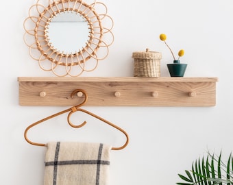 May include: A wooden shelf with four hooks, a woven rattan mirror, a small woven basket, and a small green vase with dried flowers. A brown woven hanger hangs from one of the hooks.