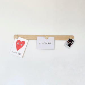 May include: A wooden wall-mounted photo display with three wooden knobs. The display has three cards attached to it. One card has a red heart with a smiley face and the text "I LOVE YOU!" The second card has the text "you are the best". The third card is a Polaroid photo of two people.