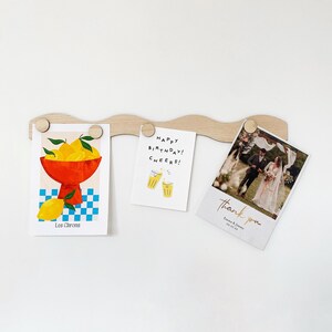 May include: A wooden wall-mounted photo display with three cards hanging from it. The cards feature a lemon illustration, a birthday greeting with a toast, and a wedding thank you card.