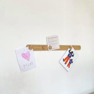 May include: A wooden wall-mounted shelf with four knobs, holding two pieces of paper. One piece of paper has a pink heart and the words "I love Mum". The other piece of paper has a drawing of two people in orange trousers and blue shirts with the word "Mama" on them. The shelf has a small card attached to it that says "Double Double Baked".