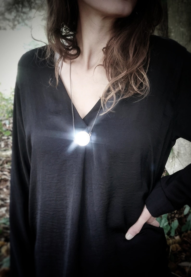 K&ouml;nnte beinhalten: Eine Frau tr&auml;gt ein schwarzes Shirt mit einer silbernen Kettenkette und einem runden, wei&szlig;en Anh&auml;nger. Der Anh&auml;nger leuchtet.
