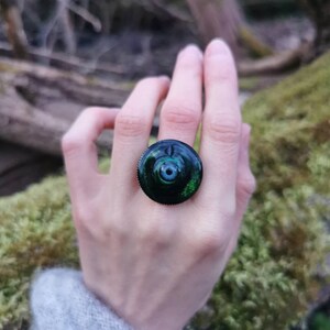 以下が含まれることがあります： 緑と青のガラス製のカボションが付いた黒い指輪。カボションは渦巻き模様で、中心は暗い。