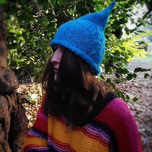 Könnte beinhalten: Eine Person trägt eine leuchtend blaue Strickmütze mit einer spitzen Spitze und einen bunten gestreiften Pullover.