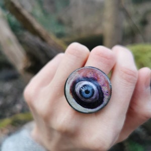 May include: A ring with a large, round, multi-colored eye design. The eye is surrounded by a silver-colored ring and has a dark blue center with a black pupil.