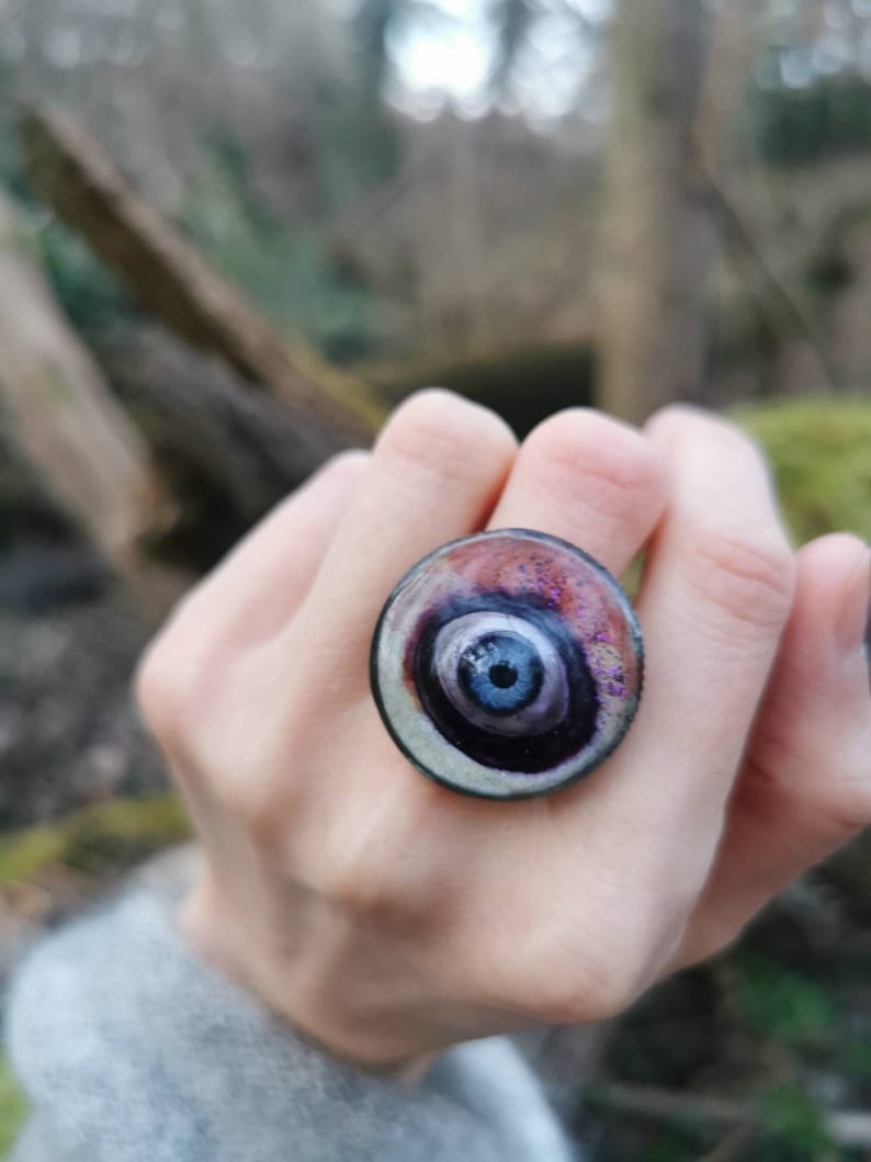 May include: A ring with a large, round, multi-colored eye design. The eye is surrounded by a silver-colored ring and has a dark blue center with a black pupil.