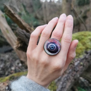 May include: A large, round ring with a swirling, multi-colored design. The ring is made of metal and has a dark blue center with concentric circles of purple, pink, and white.