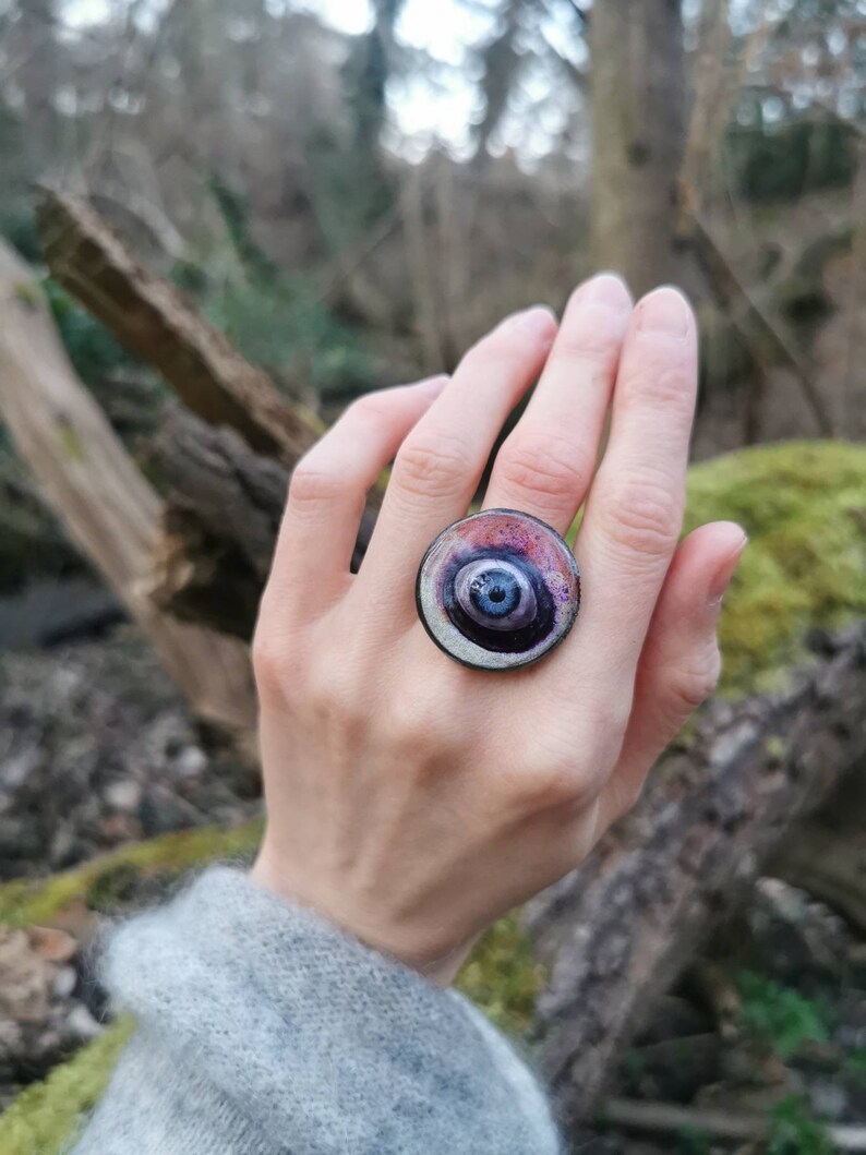May include: A large, round ring with a swirling, multi-colored design. The ring is made of metal and has a dark blue center with concentric circles of purple, pink, and white.
