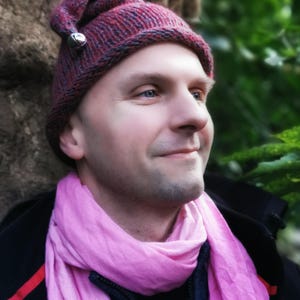 Puede incluir: Una persona con una bufanda rosa y un gorro de punto rojo y gris con una campana en la parte superior. El gorro tiene una punta puntiaguda.