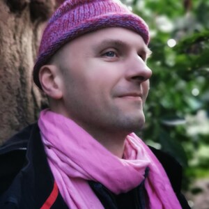Könnte beinhalten: Ein Mann trägt eine rosa Strickmütze und einen rosa Schal. Die Mütze hat eine spitze Spitze und der Schal ist um seinen Hals gewickelt.