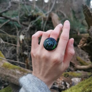以下が含まれることがあります： 銀色のベゼルにセットされた丸い緑と青の石が付いた黒いリング。リングは人の指にあります。