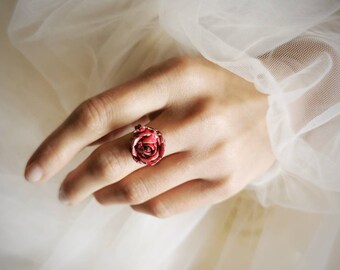 May include: A red rose ring with a gold band. The ring is worn on a hand with white fabric in the background.