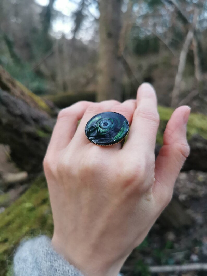 以下が含まれることがあります： 緑と黒のデザインが施された黒い指輪。指輪は丸く、金色のバンドが付いています。