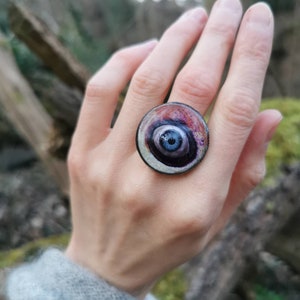 May include: A round, silver ring with a black and purple eye design. The eye has a blue center and white swirls.