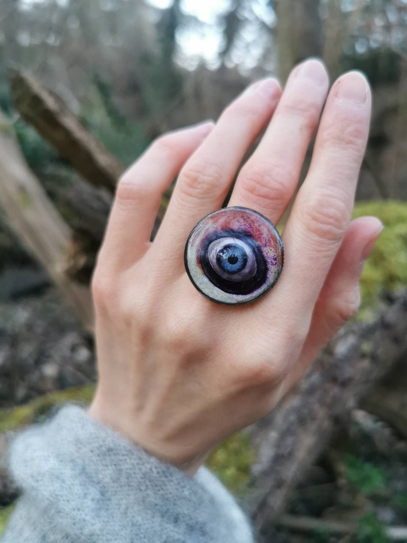 May include: A round, silver ring with a black and purple eye design. The eye has a blue center and white swirls.