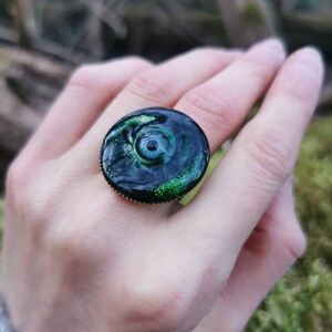 以下が含まれることがあります： 緑と青の目のデザインが施された黒い指輪。目は渦巻く緑色の模様に囲まれ、黒い瞳孔があります。